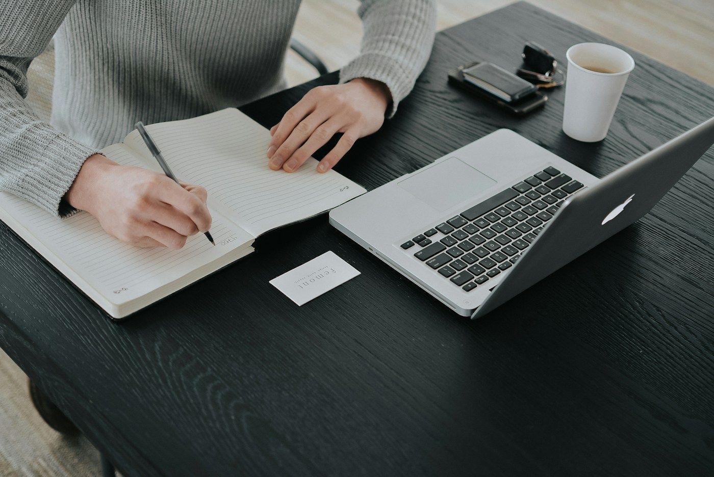 A person writing in a lined notebook beside a laptop and mug on a dark desk