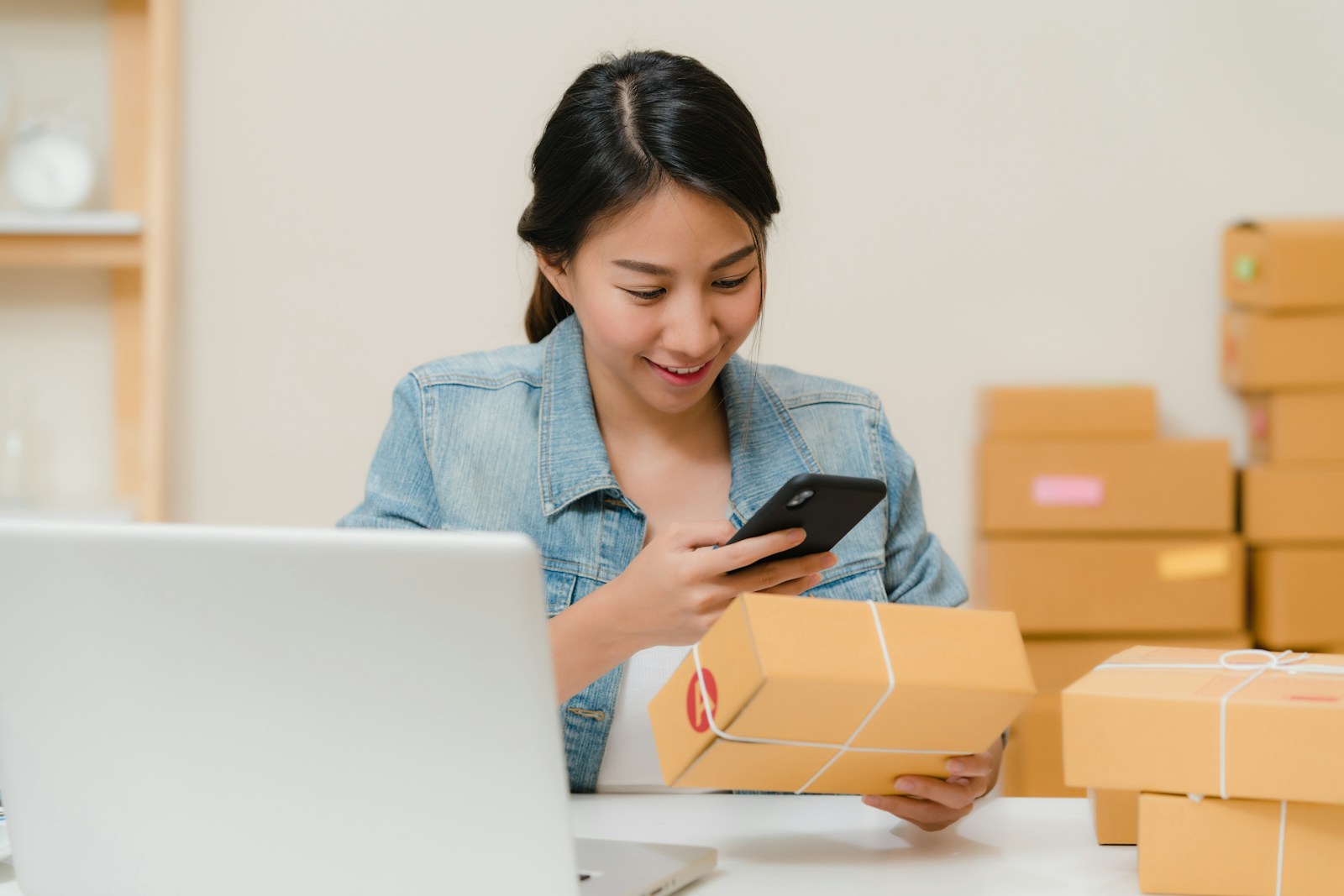 A woman checking a parcel with her phone beside a laptop and stacked boxes