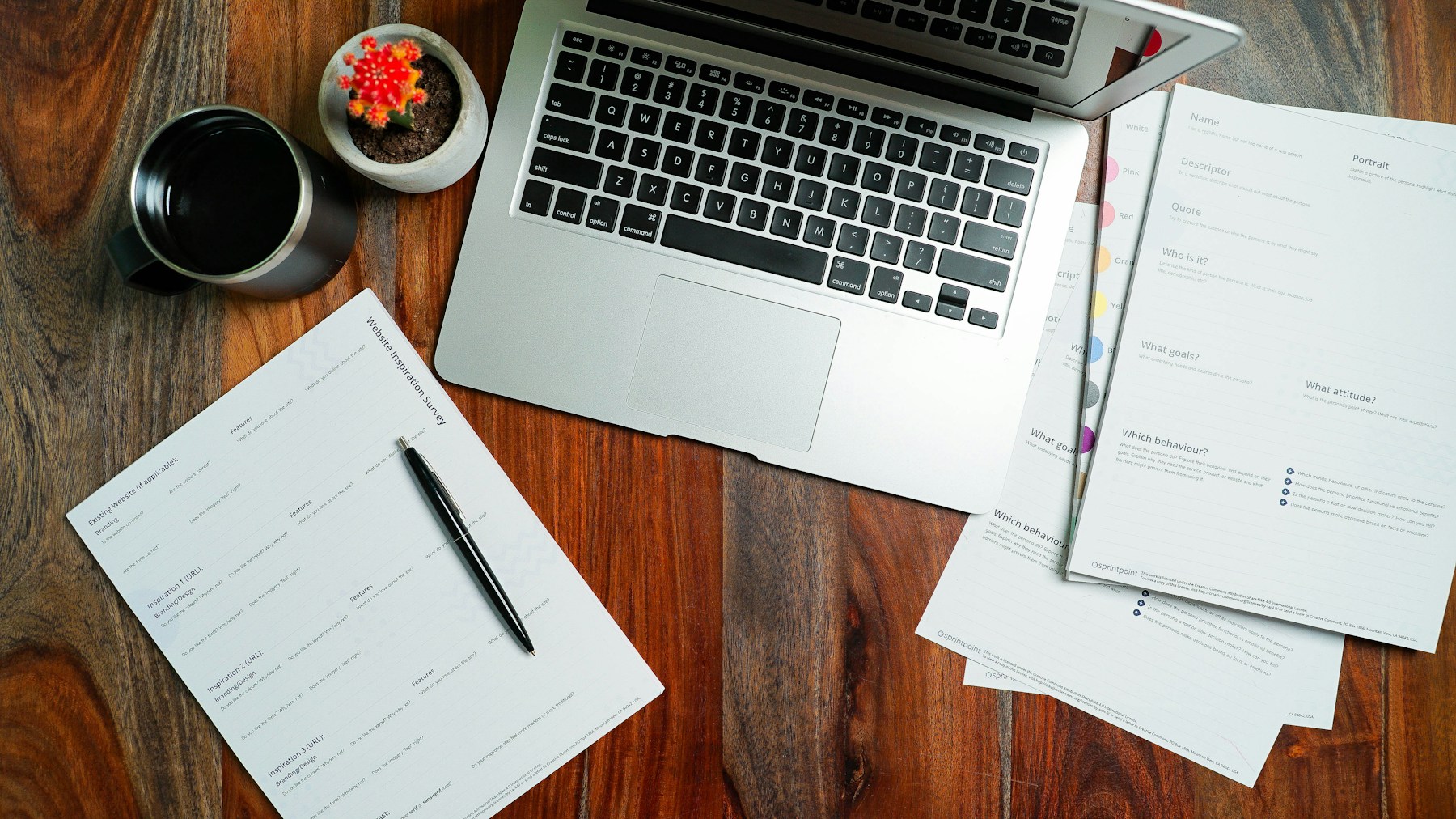 A laptop, printed worksheets, a pen and a mug arranged on a wooden desk